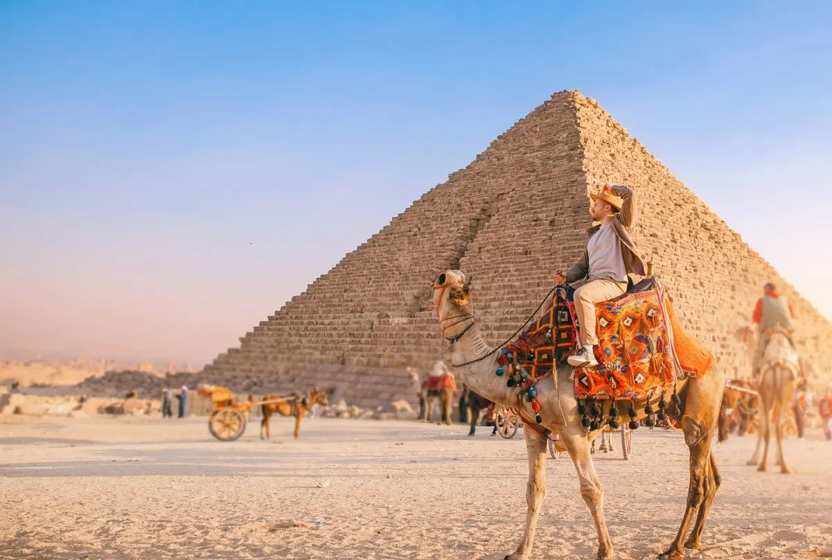 Happy Tourist man with hat riding on camel background pyramid of Egyptian Giza, sun light Cairo, Egypt.