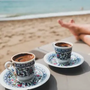 Luxury beach vacation for couple in Turkey. Two cups of Turkish coffee, female legs, sand and Mediterranean sea in background. Romantic holiday getaway for two. Summer day or morning at Antalya coast