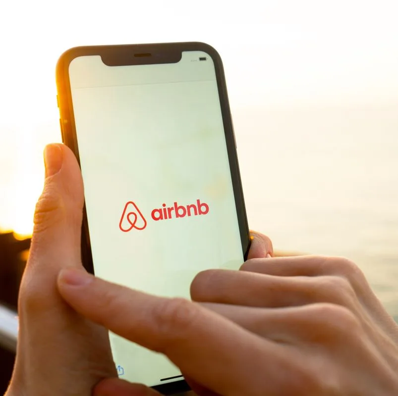 Womans hands holding smartphone displaying airbnb with sunset in the background
