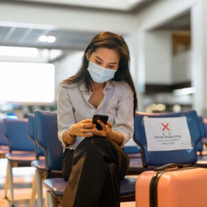 Young Female Traveler Wearing A Face Mask While Checking Her Phone At The Airport