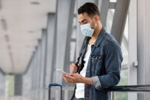 Young Male Traveler Wearing A Face Mask As He Checks His Phone At The Airport