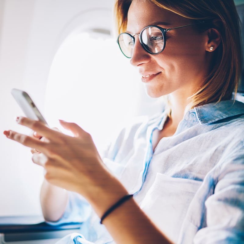Attractive female passenger on airplane reads news from network via smartphone and in-flight wifi, young woman traveling by plane in first class sending message on phone connecting wirelessly on phone