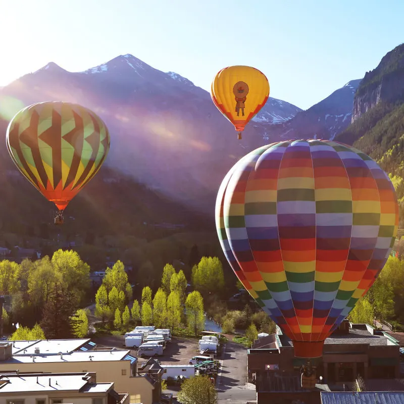 colorful hot air baloons lift up over Telluride in summertime with green trees and mountains in the distance