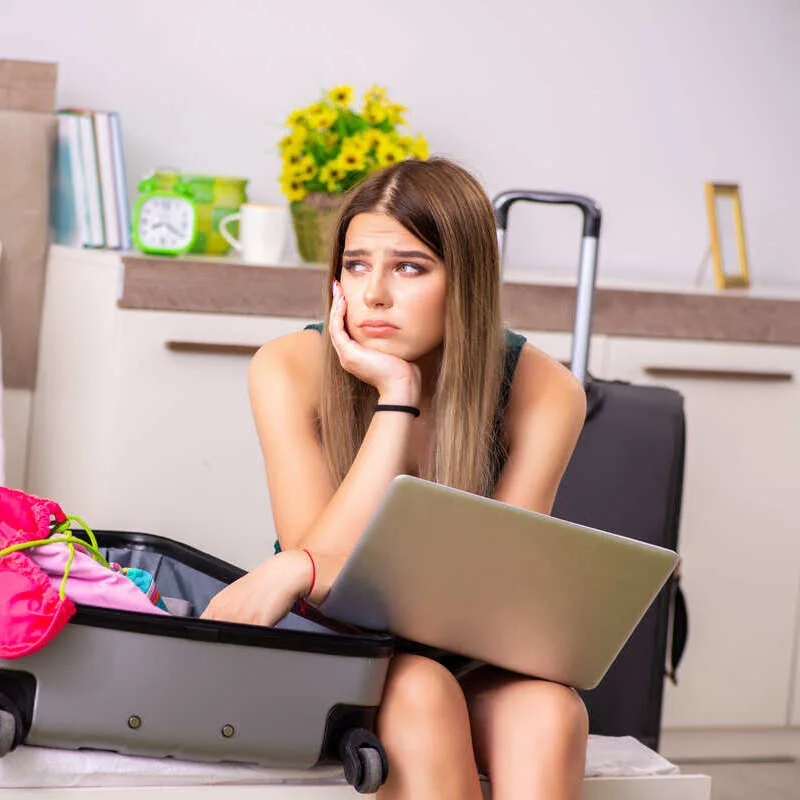 Young woman looking sad or sad sitting in her own with an open luggage on her bed and an open computer, laptop, travel chaos concept