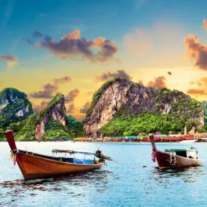 two wooden boats sit in the blue waters of phuket with tree covered rock formations in the distance under a blue and orange sky