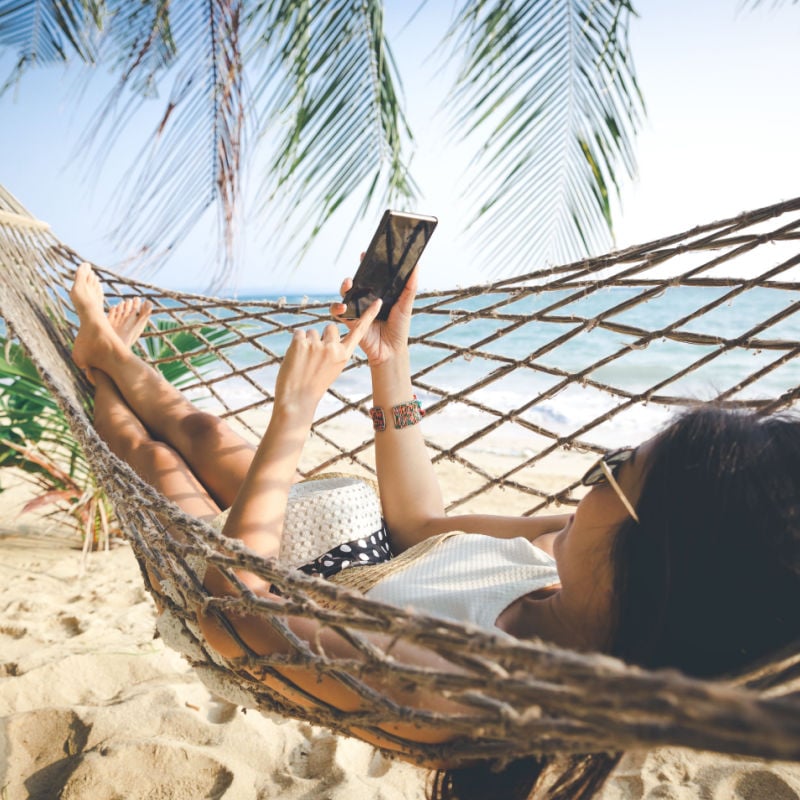 summertime holidays idea, Pleased girl with white bikini, cap and sunglass using mobile in hammock on exotic beach at sunset, Koh mak, Thailand