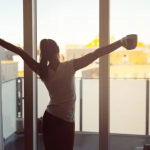 Woman stretching while holding coffee early in the morning while looking out of window