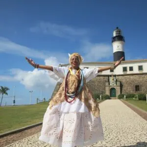 African Brazilian Woman Wearing An Ethnic Garment As She Stands Smiling With Her Arms Open In Front Of A Historic Fort In Salvador De Bahia, Northeastern Brazil