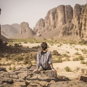Female Tourist Sat On A Rock As She Admires The Rugged Landscape Of Djanet In Southern Algeria, North Africa