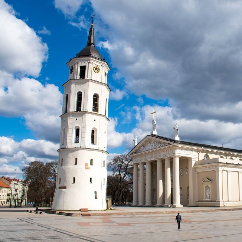 Manlig turist som går på ett tomt huvudtorg i Gamla stan i Vilnius, där ett vitt medeltida torn och katedralen ligger, Vilnius, Litauen, ett land i östra Europa vid den baltiska kusten