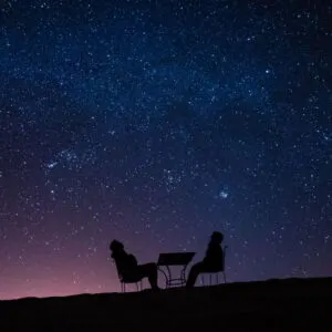 young couple sitting at a table on a desert dune while talking, relaxing and observing the stars and the milky way above them