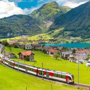 Traditional Red Tourist Train Traveling Through A Picturesque Region Of Switzerland, By The Alps, Where A Small Swiss Village And Alpine Lake Is Located, Central Europe
