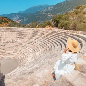 Elegant and charming woman in white dress and hat explores ancient landmark and ruins of Greek amphitheater in Kas resort, Turkey