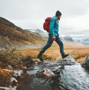 hiker crossing a stream with white clouds and mountains in the back