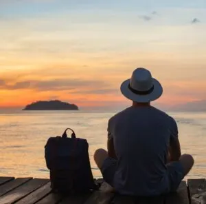 man with hat sitting on a dock with his backpack looking at the sunset