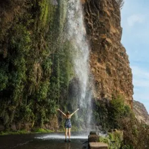 A Young Woman Raising Her Arms As She Admires A Waterfall Called Cascata Dos Anjos In Madeira, Portugal
