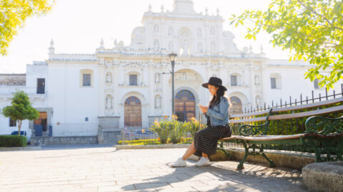 Young Hispanic woman sitting watching her phone in front of the San Jose cathedral in Antigua Guatemala-tourist traveling in colonial city-woman in the central park of Antigua Guatemala