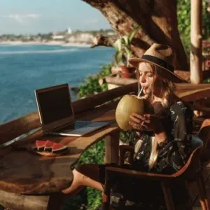 Young Female Digital Nomad Sipping On A Coconut As She Works From Her Computer In A Tropical Location In Bali, Indonesia, Southeast Asia