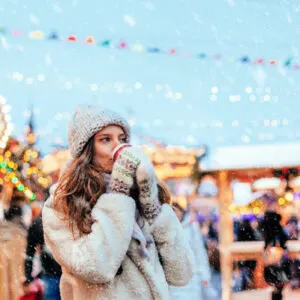 Girl drinking hot coffee while walking in Christmas market decorated with holiday lights in the evening. Feeling happy in big city