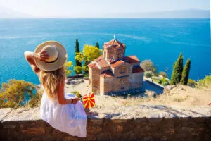 Young Woman Wearing A Summery Dress As She Waves A Small Macedonian Flag While Admiring A Byzantine Church Facing Lake Ohrid, North Macedonia, South Eastern Europe