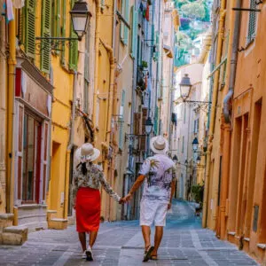 Young Couple Walking Down A Narrow Alley Lined By Pastel Colored Buildings In Menton, French Riviera, Cote d'Azur, Southern France, Southern Europe