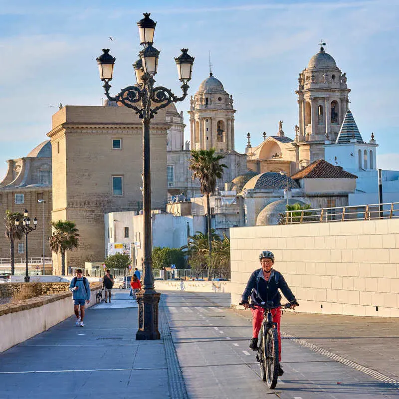 People Biking Along The Promenade By Cadiz Cathedral, Spain