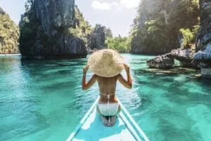 Woman in sun hat on the bow of a boat in a beautiful turquoise tropical lagoon surrounded by cliffs in Philippines