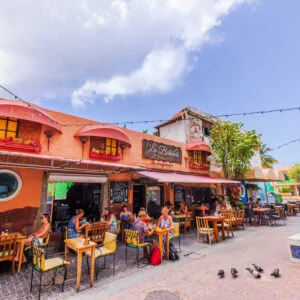 Willemstad street with outdoor cafe, colonial buildings, people dining, wooden tables, tropical plants and pigeons on pavement. Willemstad. Curacao