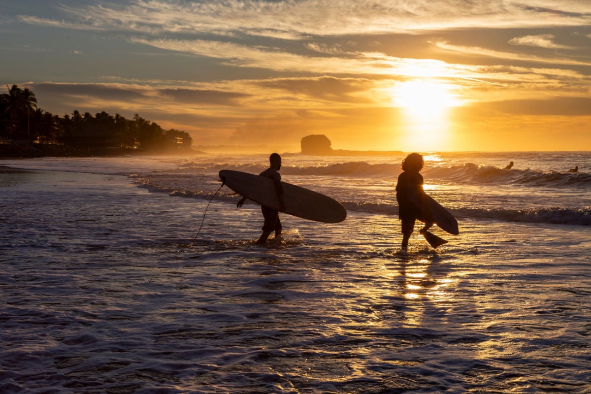 Two surfers at Playa El Tunco, El Salvador, walk into the waves against a vibrant sunrise at this popular surfing beach. One man has swimming fins and a bodyboard. The iconic rock pile is in the distance.