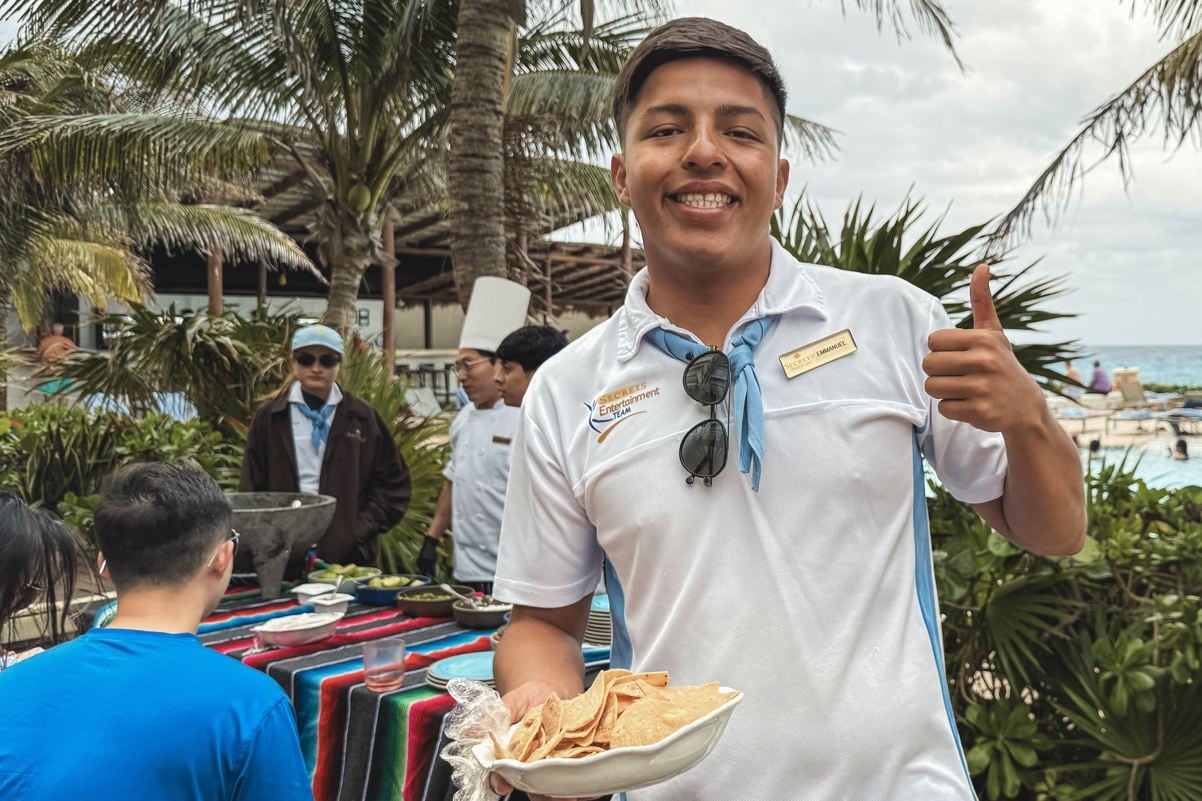 A smiling staff member at an all inclusive resort in Mexico holding a bowl of chips and giving a thumbs up, representing the hospitality workers who serve US travelers.
