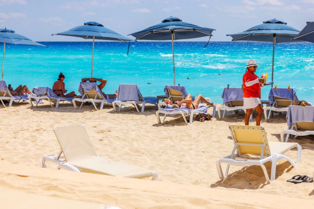 Caribbean-beach-with-tourists-relaxing-on-sun-loungers-under-blue-umbrellas-while-waiter-in-serves-drinks-against-turquoise-sea-waves.-Mexico.-Cancun