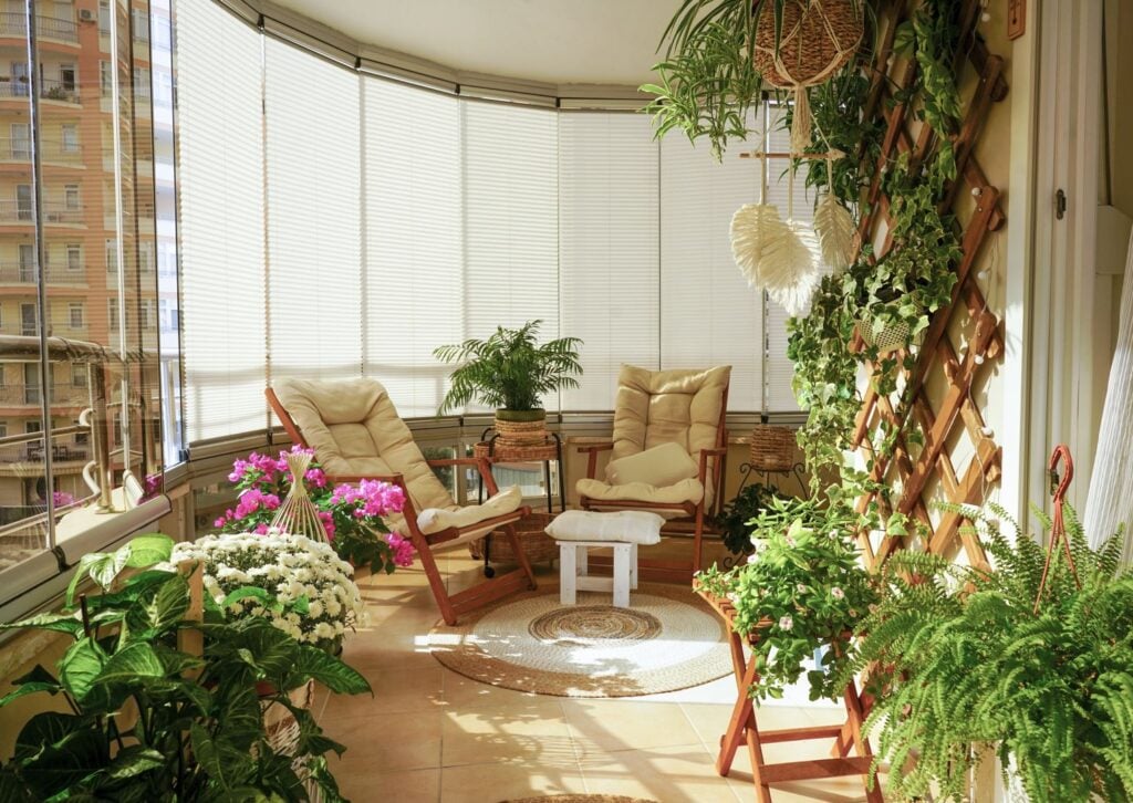Bright, cozy sunroom balcony decorated with lush potted plants, wicker accents, and folding chairs