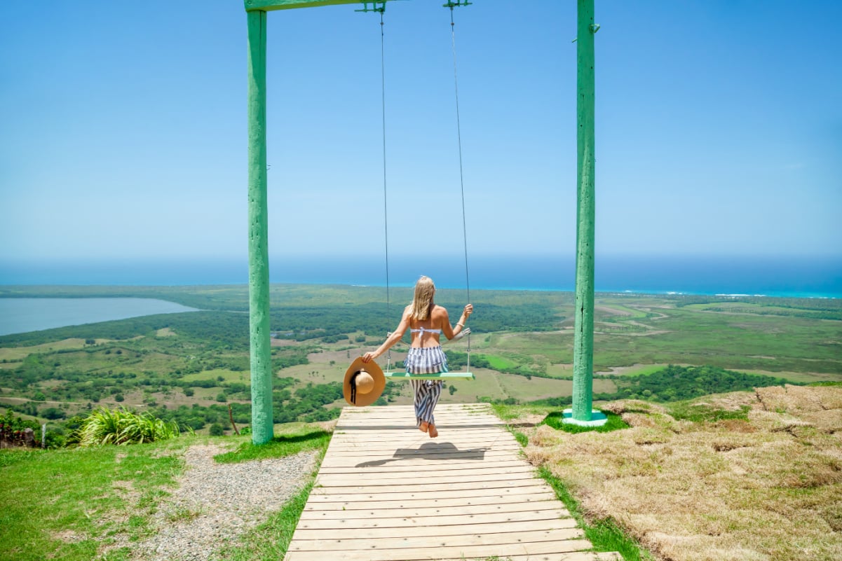 Woman swinging on swings over the green mountains with ocean sea caribbean tropical landscape and horizon line with blue sky. Montana Redonda, Dominican republic. Freedom concept. Touristic place