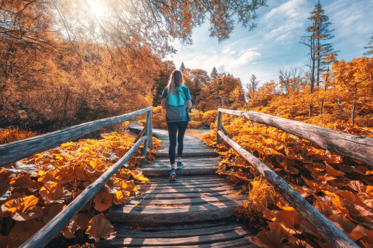 Young woman with backpack walking along a wooden path near stunning waterfalls, orange trees in Plitvice Lakes national park, Croatia at sunset in autumn. Sporty girl in forest in fall