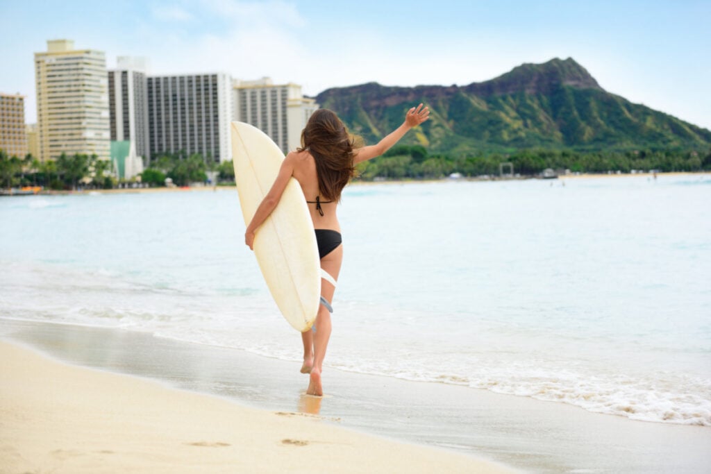 Beach fun - Happy freedom surfer woman going surfing in Waikiki beach, Honolulu city, Oahu, Hawaii, USA. Hawaiian surf girl heading for waves with surfboard living healthy active lifestyle, rear view.